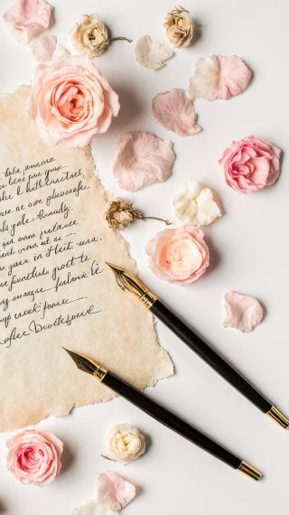Photograph of a vintage paper letter with a fountain pen, rose petals, and a box of chocolates in soft pastel colors Wallpaper